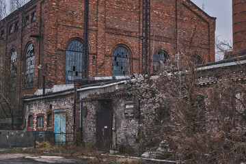 Exterior view of a massive abandoned red brick factory with broken arched windows and crumbling stone walls. A derelict industrial site showing urban decay, weathered facades, and overgrown weeds.
