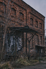 Abandoned red brick industrial building with arched windows and rusted metal awning. Gloomy atmosphere of a derelict warehouse with graffiti, overgrown grass, and urban decay under a cloudy sky.