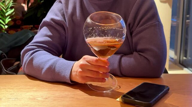 Hand Holding Wine Glass Amber Liquid, Phone On Wooden Table, Sweater Sleeve Visible, Warm Bistro Lighting And Soft Reflections On Glass. Freelancer Taking Quiet AfterWork Break, Checking