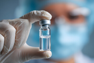 Medical professional wearing protective gloves and mask holding a small transparent vial with clear liquid in focus for scientific or healthcare purposes