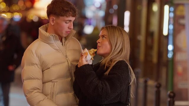A couple enjoys street food in Paris France at night The woman takes a bite of a sandwich while the man looks on The scene captures a romantic date in the city with a bokeh lights background.