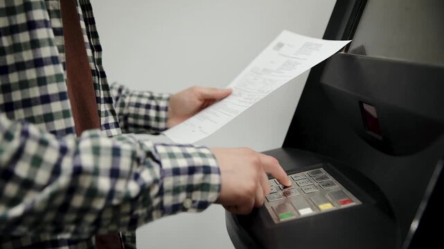 Man standing at an atm, focused on a document in his hand as he enters information on the keypad to complete a banking transaction. Transferring funds to a bank account using a bank terminal