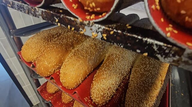 Camera moves upward along bakery racks filled with freshly baked bread rolls topped with sesame seeds.
