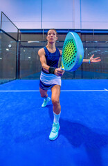 A determined and focused female padel player in a perfect waiting position, awaiting her opponent's ball to return it on a blue indoor court.