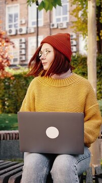 Young female sitting on bench in city park, working online on laptop. Cruiser bike nearby. Autumn sunny day