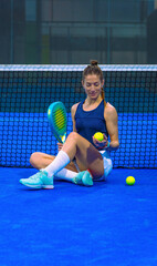 Female padel player sitting on a blue court, in front of the net, smiling while holding a racket and looking at a padel ball, motivating herself for the match.