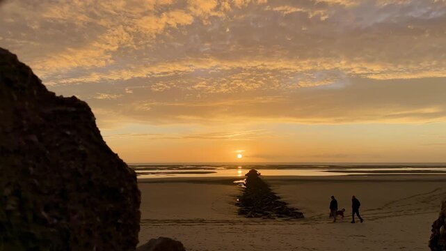 Time-lapse sunset over Authie Bay in Berck, France, golden clouds racing above low tide sand flats, rocky groyne to the horizon with walkers and dog silhouettes