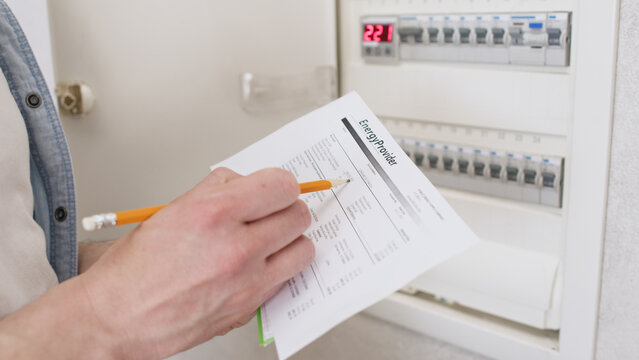 A close-up of a man holding a bill for electricity and underlining the main amount with a pencil; in the background is an electrical panel with switches for the apartment.