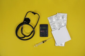 stethoscope, syringe, calculator, and medicine blister packs on an yellow background. Illustrates medical expenses, health budgeting, insurance, and financial planning in healthcare.