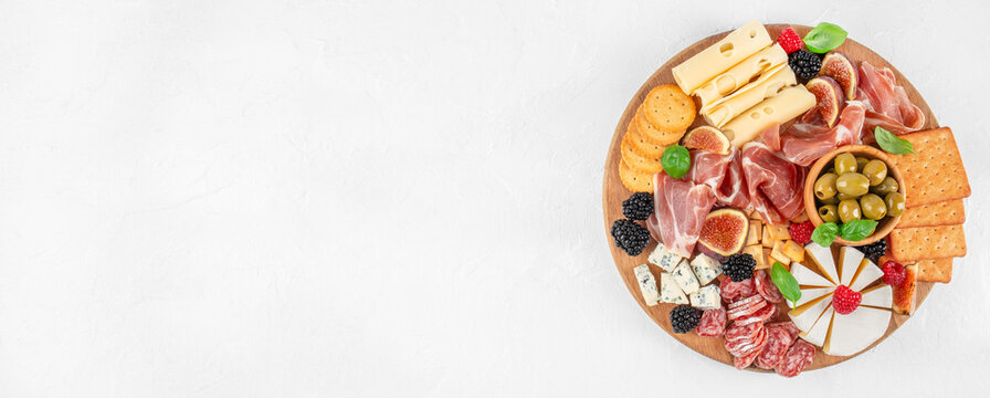 Top view of gourmet charcuterie board with cheese, prosciutto, olives, crackers and fresh berries on white background with copy space