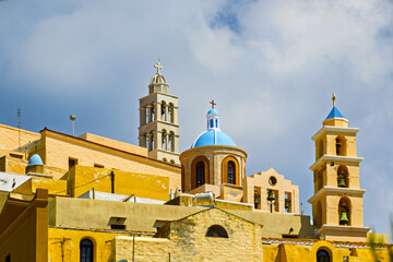 view of Ano Syros, Greece, showing a densely built hillside town with traditional Cycladic architecture. The buildings feature pastel facades with tiled roofs cascading down the slope in layered forma