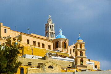 view of Ano Syros, Greece, showing a densely built hillside town with traditional Cycladic architecture. The buildings feature pastel facades with tiled roofs cascading down the slope in layered forma