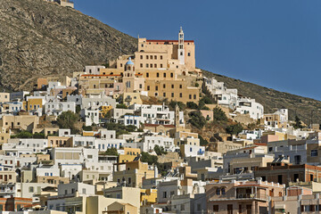 view of Ano Syros, Greece, showing a densely built hillside town with traditional Cycladic architecture. The buildings feature pastel facades with tiled roofs cascading down the slope in layered forma