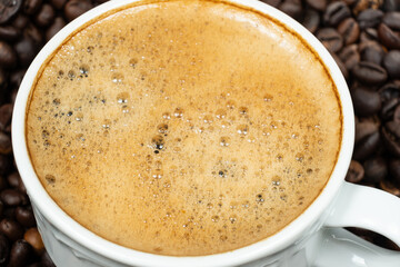 Aromatic coffee with rich bubbly foam in a cup surrounded by roasted beans.
