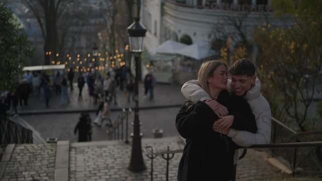 A romantic couple embraces in Montmartre Paris France The woman kisses the man on the cheek The background is blurred showing the Sacre-Coeur basilica and a bustling street scene.
