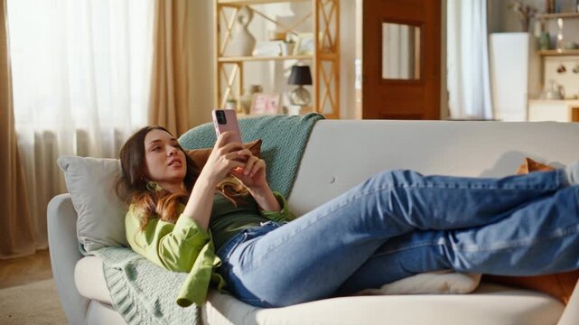 Relaxed girl watching smartphone screen lying comfortable couch. Smiling young