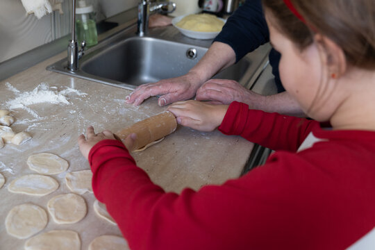 Grandpa guides granddaughter's hands on rolling pin over dough near kitchen sink, selective focus on joyful multi-generational cooking at home during holiday dumpling making