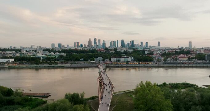 Aerial view of Warsaw skyline over touristic Pedestrian and Bicycle Bridge crossing the Vistula River. Urban riverfront with modern infrastructure and city landmarks. Cinematic 4K drone shot