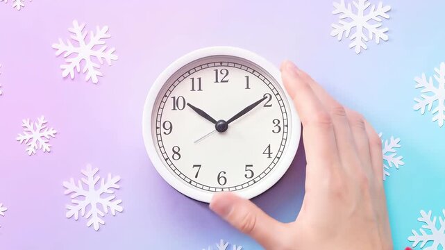 Hand holds a small round clock against a soft pastel gradient with scattered paper snowflakes. A red knit sleeve suggests winter and holiday time. The flat lay composition emphasizes seasonal timing a