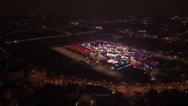 Drohnenaufnahme bei Nacht von der Theresienwiese in Muenchen. Sichtbar sind das Tollwood Winterfestival, leuchtende Zelte und der hell erleuchtete Stadtkern im Winter.