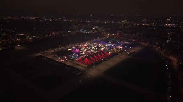 Drohnenvideo bei Nacht ueber die Theresienwiese in Muenchen. Die Luftaufnahme zeigt das Tollwood Winterfestival mit leuchtenden Zelten, Weihnachtsmarkt und heller Stadtbeleuchtung im Winter.