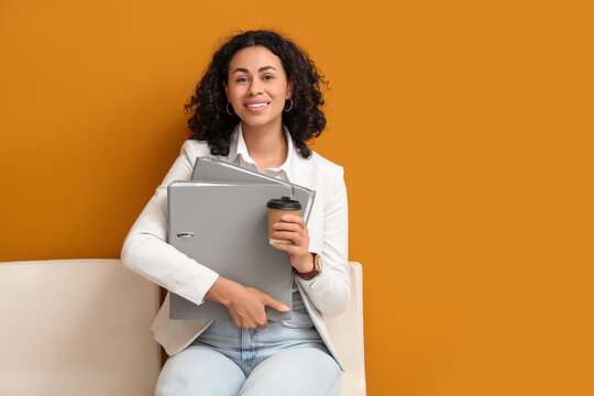 Female African-American job applicant with folders and coffee waiting for interview near orange wall