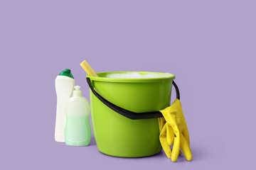 Plastic bucket with foam, sponge, rubber gloves and bottles of detergents on purple background