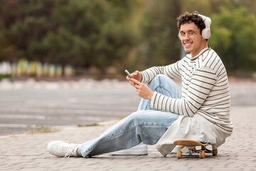 Happy young man in headphones with mobile phone sitting on skateboard outdoors