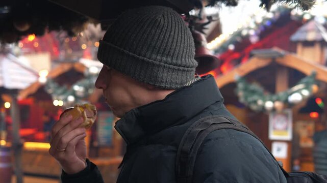 A man eats a hot dog at a Christmas market. Cozy winter atmosphere, festive lights and decorations in the background.