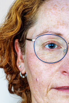 Half face close up of a mature freckled redheaded woman with a blue eye and natural skin wearing round eyeglasses.
