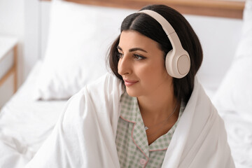 Beautiful young woman with modern headphones and blanket listening to music in bedroom