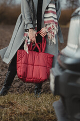Stylish woman in a gray coat and striped scarf holding a red quilted puffer bag against a dark field, vertical shot in natural light