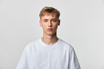 Fototapeta premium Young man in a white t shirt stands before a neutral background in a studio shot, presenting a calm expression and modern simplicity with clean lighting and minimal styling, short blond hair.