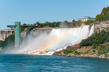 View of Niagara Falls with mist and rainbow, featuring the observation tower and river below on a clear summer day. Popular travel landmark on the US&ndash;Canada border.