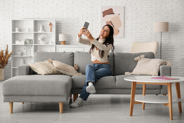 Pretty young woman with mobile phone taking selfie on sofa at home