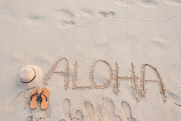 Aloha word written in beach sand with hat and sandals © Sulbha
