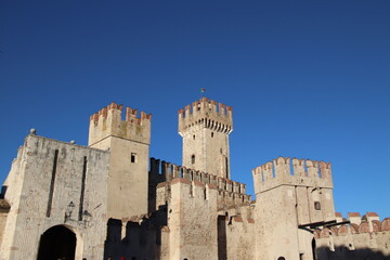Vertical perspective of the Scaligero Castle in Sirmione. This iconic 14th-century medieval fortress features distinctive swallow-tail merlons (Ghibelline style), high defensive towers, and ancient st