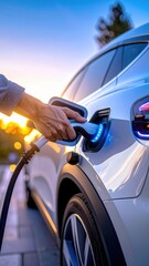 Person Charging White Electric Vehicle Outdoors During Golden Hour Sunset With Soft Bokeh Lights In Background
