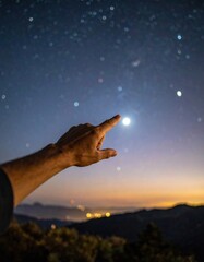 Human Hand Reaching Towards Bright Star In A Clear Night Sky Over Mountain Landscape With Distant City Lights