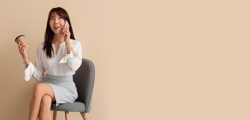 Asian businesswoman with coffee talking by mobile phone on chair against beige background with space for text