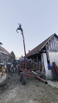Rusty wood trailer hitched to a tractor using a mechanical loading arm to precisely handle a small log in a farmyard in Central Europe.
