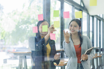 Young businesswoman creative team using post it notes in glass wall to writing strategy business plan to development grow to success.

