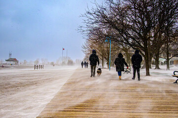 People walking along a wooden boardwalk at Kew Beach, Toronto, with blowing snow over frozen sand; benches, bare trees and a dog walker visible in winter.