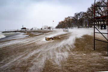 Wind streaks of drifting snow move across frozen sand at Kew Beach in Toronto, with a red lifeguard chair and the Lake Ontario shoreline visible under a grey sky.