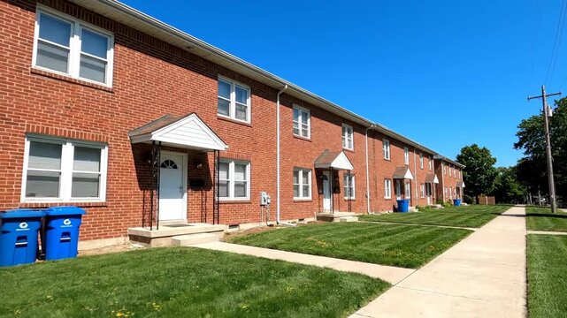Row of brick apartment buildings with green lawn under blue sky, showcasing typical residential architecture, urban neighborhood and sidewalk.
