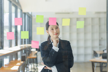 Thoughtful asian woman with hand on chin looking at sticky notes on glass wall, 
