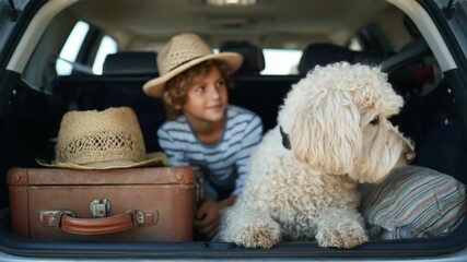 Ready for adventure: A young boy and his furry friend are packed, ready and raring to go on a vacation, sitting comfortably among luggage in car trunk. Capturing anticipation and excitement.