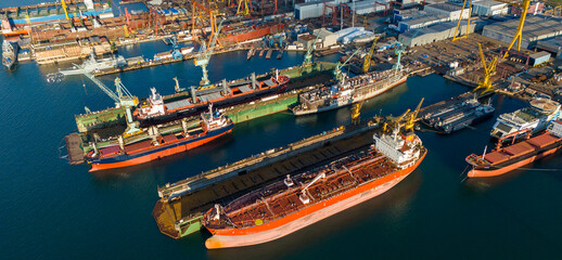 Side view drone shot of a large orange tanker ship lifted in a floating dry dock for hull inspection and painting surrounded by other docked industrial vessels © Buğra Kaan Ersoy