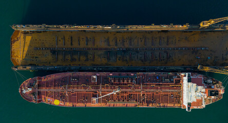Horizontal top down drone shot of a long tanker ship in a dry dock showing the complex network of pipes on the deck and the yellow walls of the floating repair dock