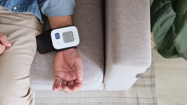 Unrecognizable man using a digital tonometer to measure his blood pressure while sitting on a sofa